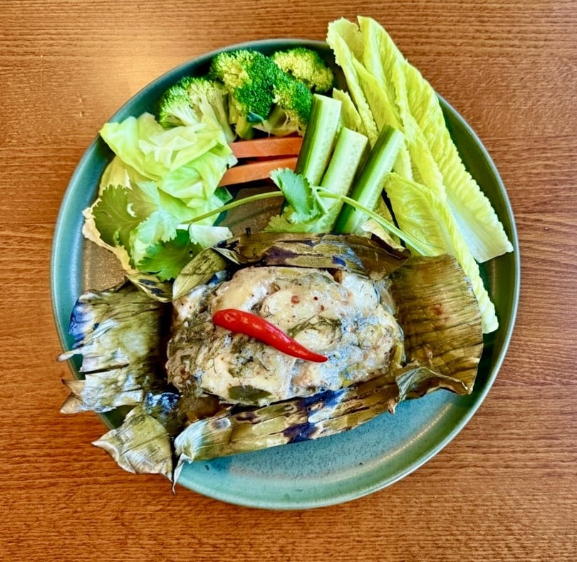 Steamed fish in banana leaves with a red chili, served with fresh vegetables on a green plate.