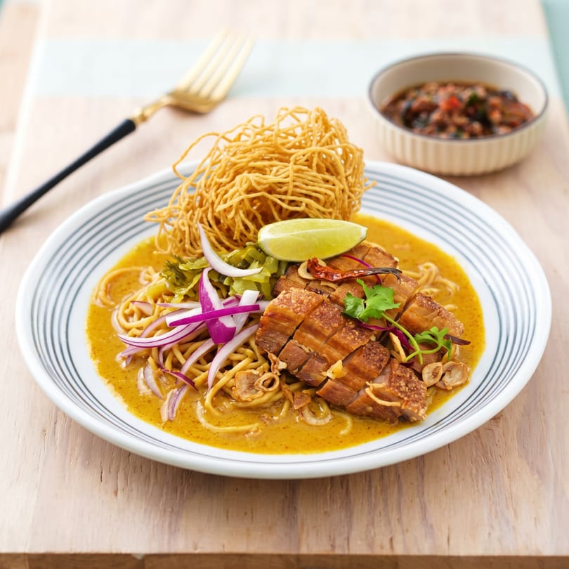 Khao Soi with crispy pork belly, red onions, lime, and crispy noodles in a bowl.