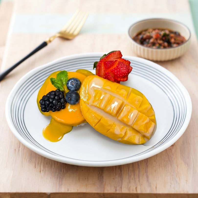 Mango sticky rice dessert with berries and mint on a white plate, with a small bowl of sauce.