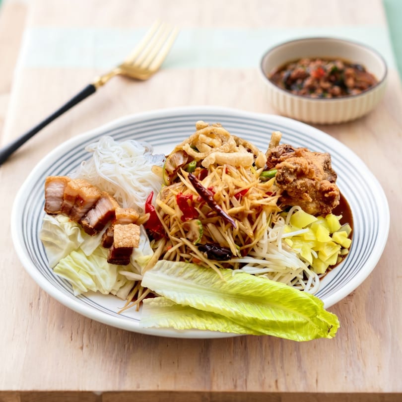 A vibrant Thai papaya salad with crispy pork belly, fried chicken, noodles, and vegetables.